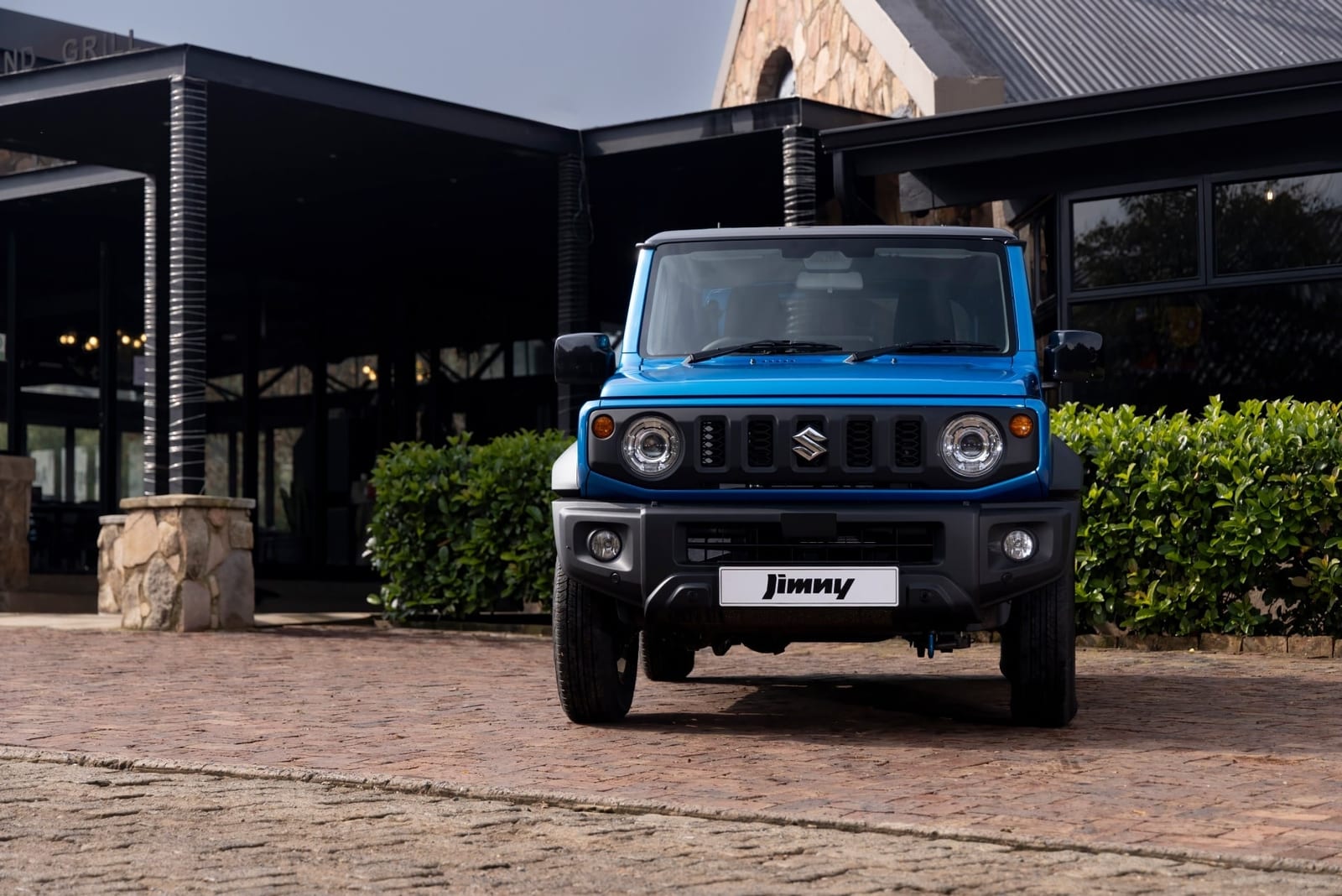 A standard blue Suzuki Jimny 3-door parked on brick paving in front of a stone and glass building