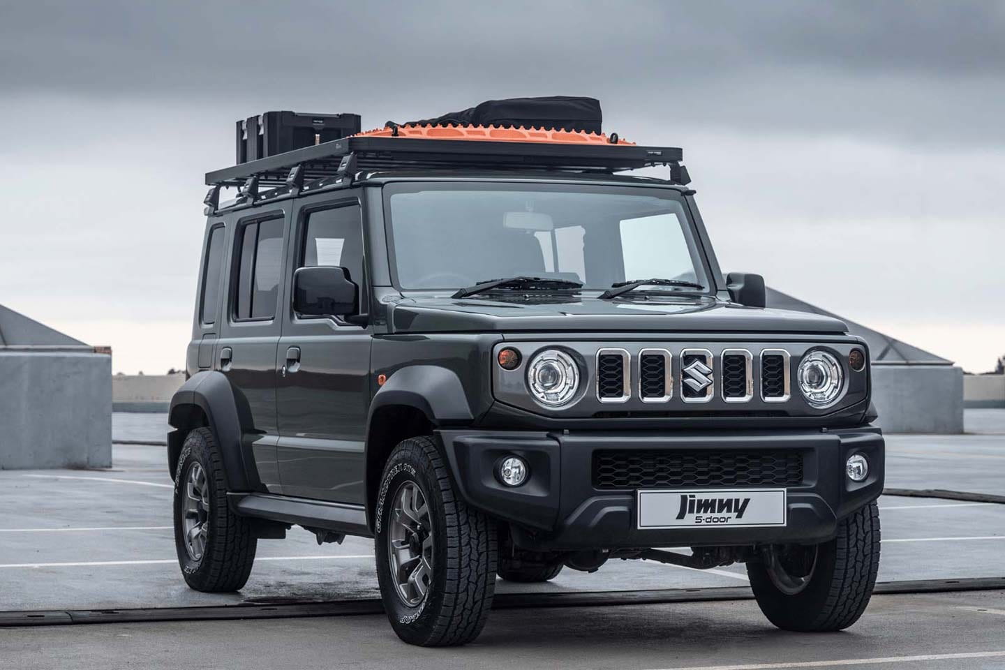 A dark grey Suzuki Jimny 5-door with a roof rack and recovery boards parked on concrete under a grey sky