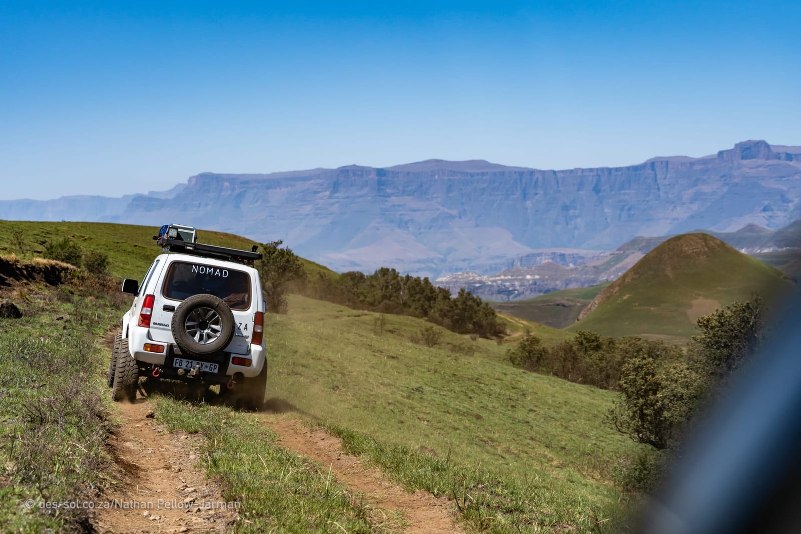 A white older Suzuki Jimny with a roof rack and rear spare wheel driving along a grassy farm track, with a long escarpment behind