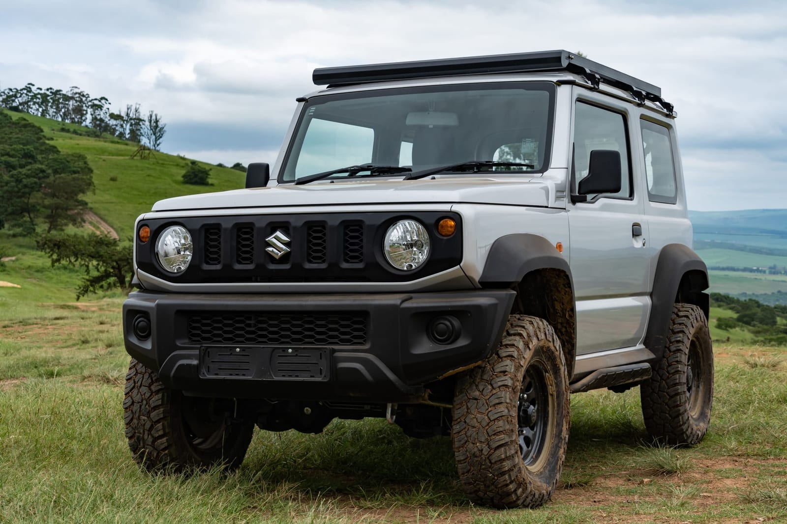 A silver current Suzuki Jimny 3-door with a flat roof rack, mud-terrain tyres on black steel wheels and a mild lift, parked on green grass
