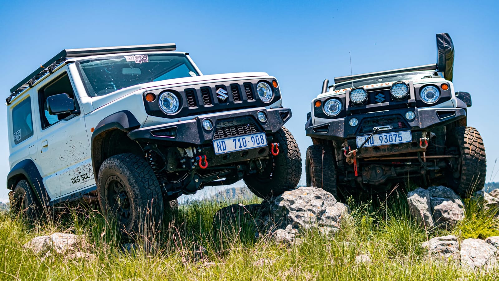 Two current Suzuki Jimny 3-doors with steel front bumpers, rock sliders and roof racks, parked on rocks and grass under a blue sky