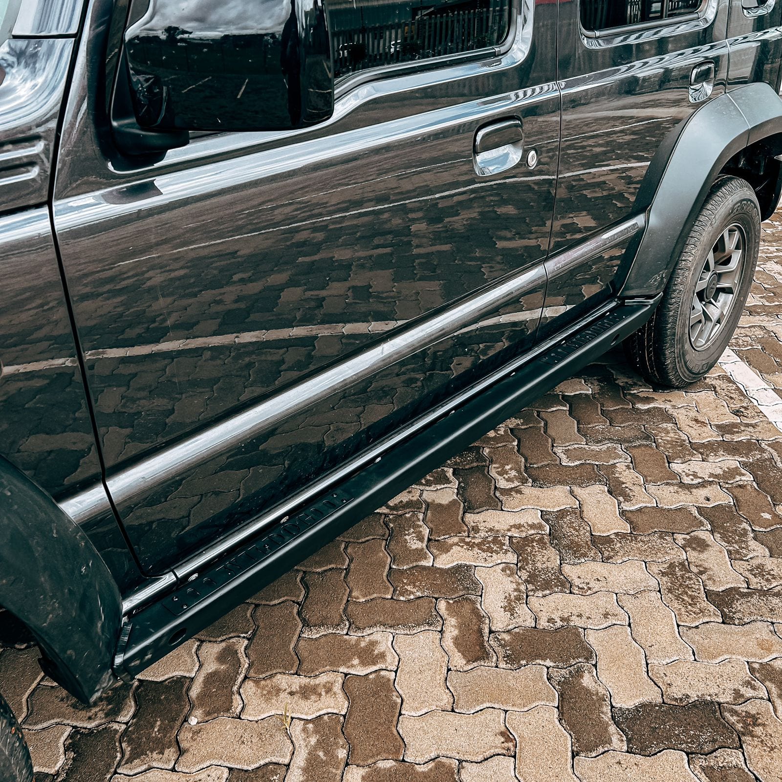 A dark grey Suzuki Jimny 5-door seen side on, with black steel rock sliders running along the sill between the wheels