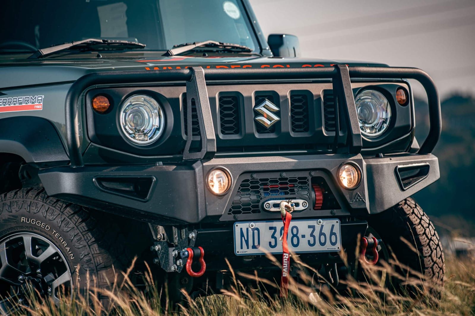 The front of a black Suzuki Jimny with a steel hoop bumper, a winch with a red hook, red shackles and round spotlights, standing in long grass
