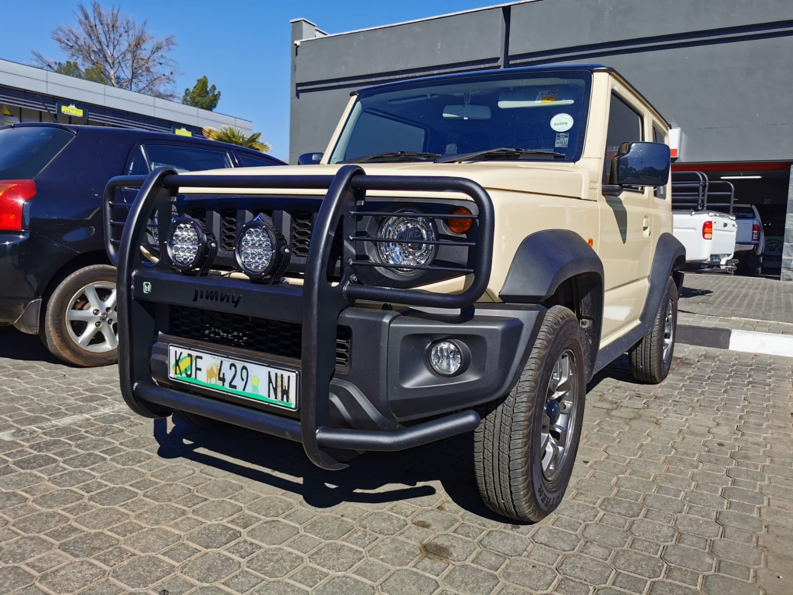 A beige Suzuki Jimny 3-door with a black wrap-around bull bar and two spotlights fitted over the standard bumper