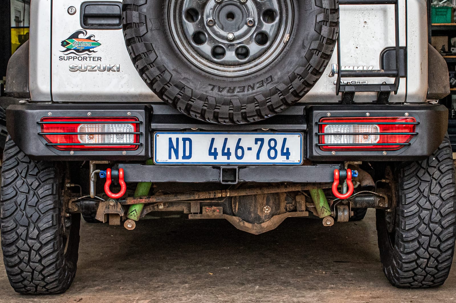 The back of a white Suzuki Jimny 3-door with a steel rear bumper, a tow hitch, red shackles and the rear lights set into the bumper, with a large spare wheel on the back door