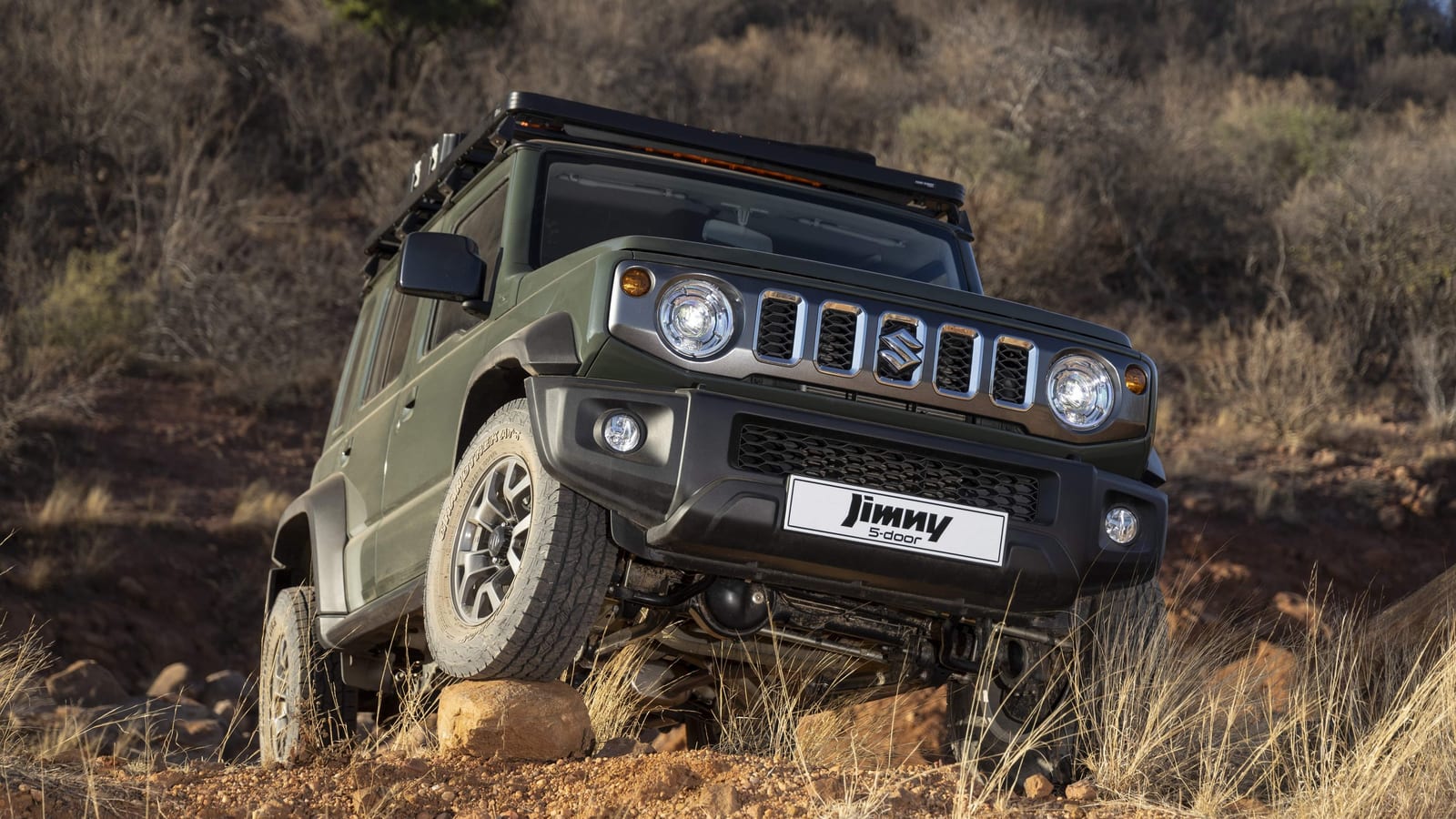A green Suzuki Jimny 5-door with a full-length roof rack, one front wheel up on a rock on a dry bushveld slope