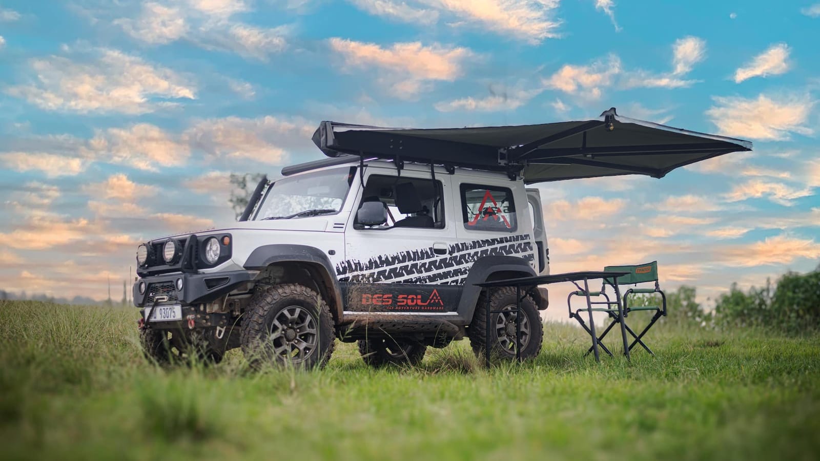 A white current Suzuki Jimny 3-door on grass with a side awning rolled out from its roof rack, a camp chair and table beside it