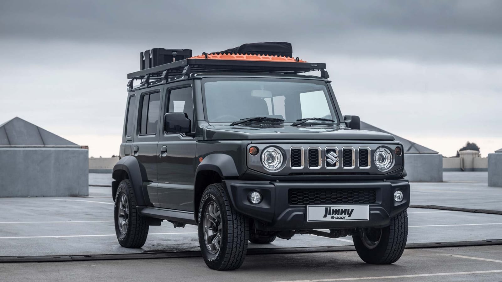 A dark grey Suzuki Jimny 5-door on a rooftop car park with two black boxes, orange recovery tracks and a bag on its roof rack