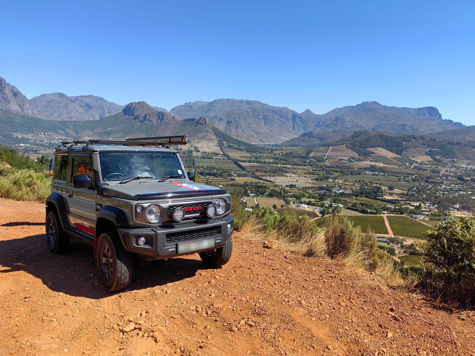 A grey current Suzuki Jimny 3-door with two load bars on the roof, parked on a red gravel lookout above a valley of vineyards and mountains