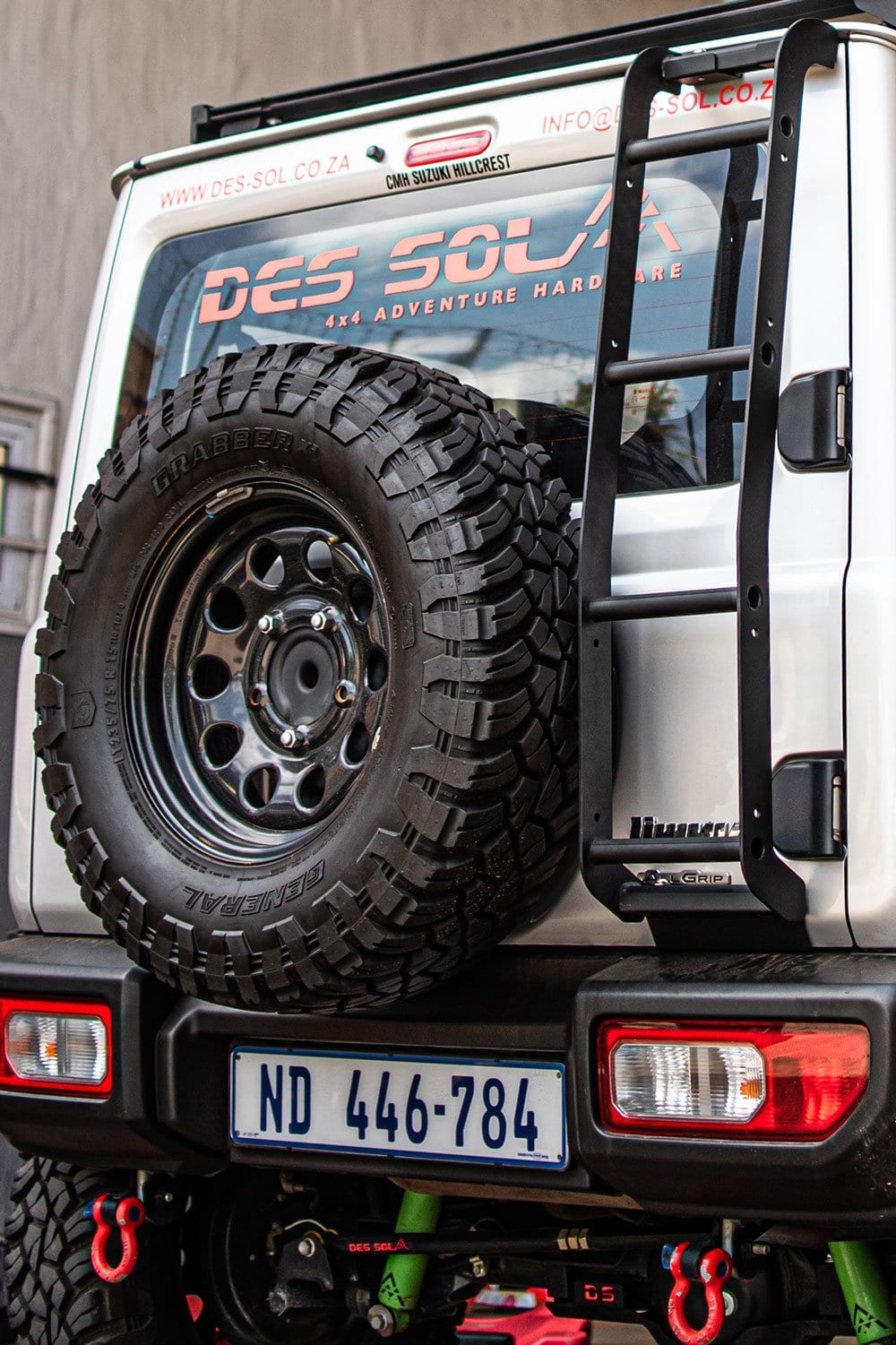 The back of a silver current Suzuki Jimny 3-door with a black ladder fixed beside the spare wheel on the side-hinged back door