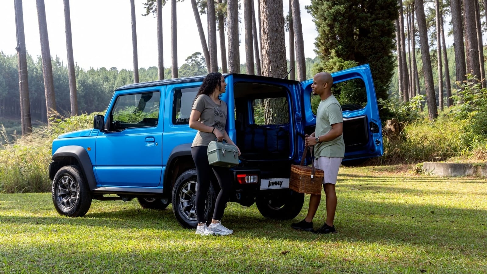 A blue Suzuki Jimny 3-door with its back door open in a pine plantation, a couple standing beside it with a picnic basket and a cooler bag