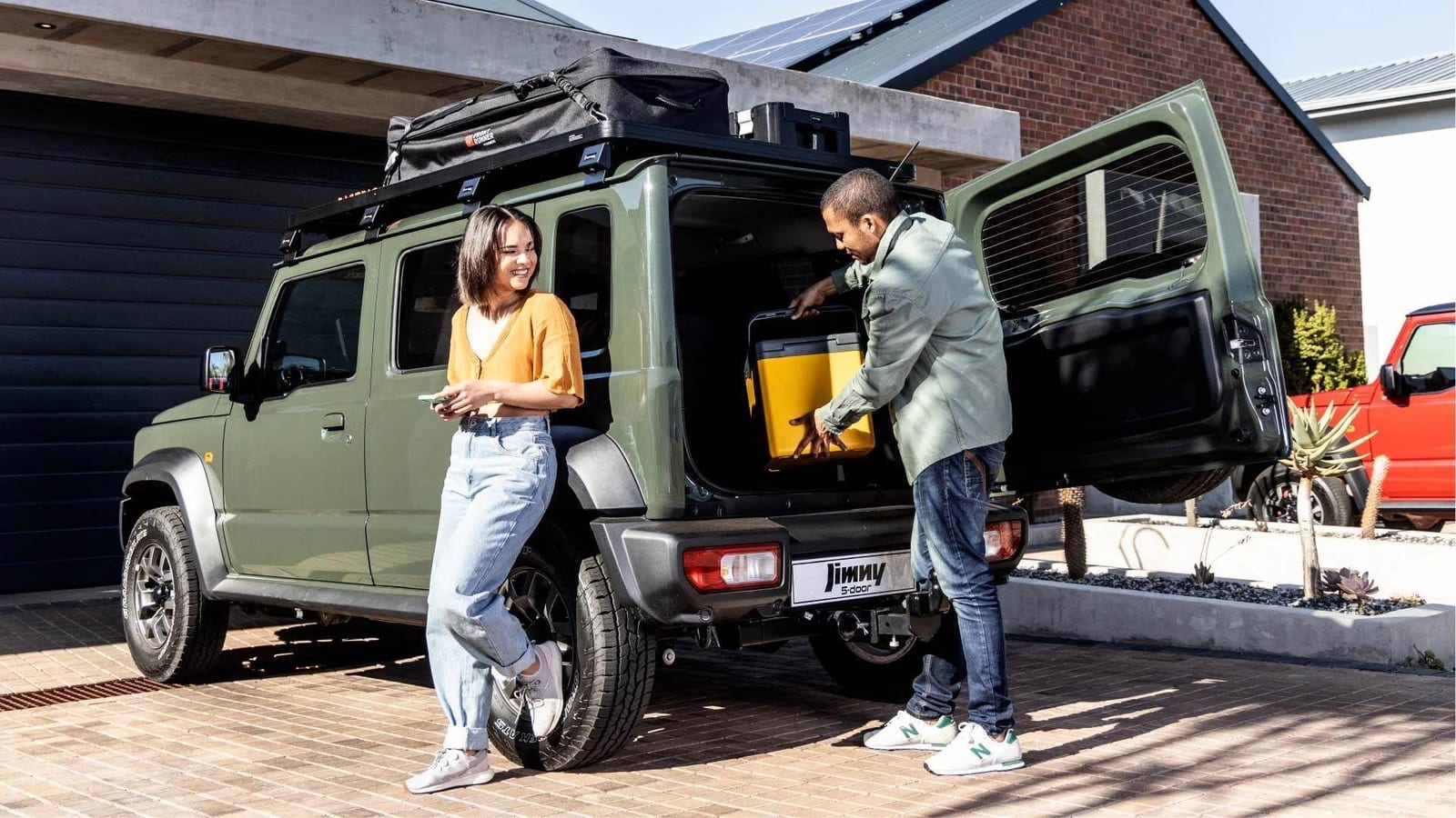 A green Suzuki Jimny 5-door in a driveway with its back door open, a man lifting a yellow cooler box into the boot while a woman watches