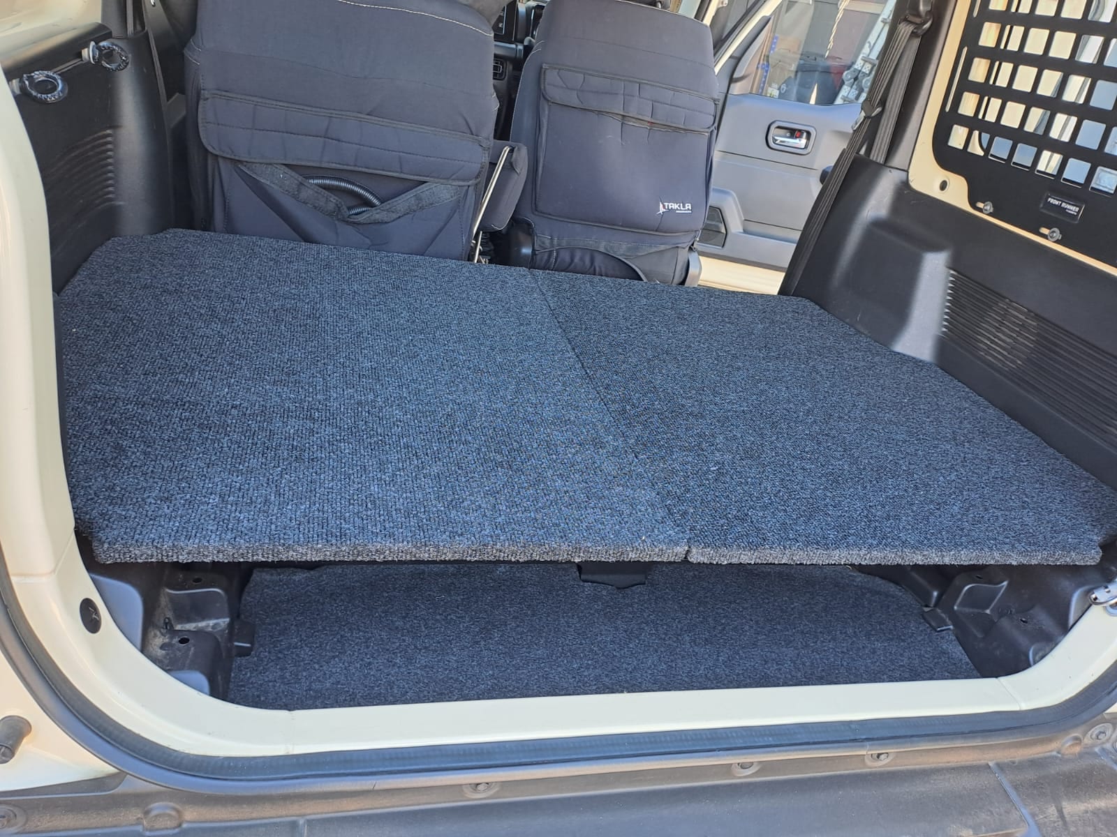 A grey carpeted flat deck in the boot of a current Suzuki Jimny 3-door, with the rear seats folded forward and a Molle panel on the side window