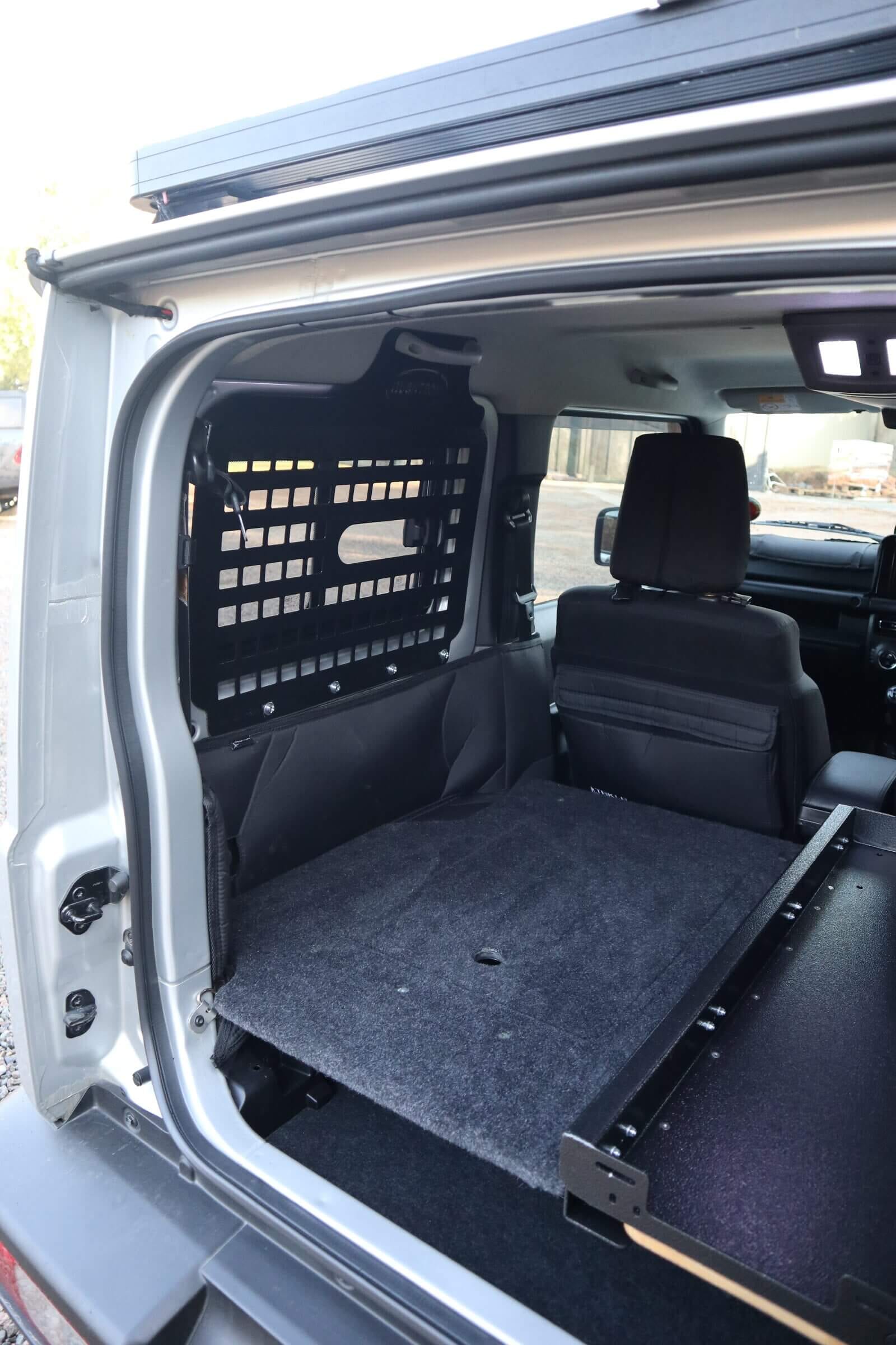 Inside a current Suzuki Jimny 3-door with the rear seats out: black Molle panels over both rear side windows and a flat carpeted floor