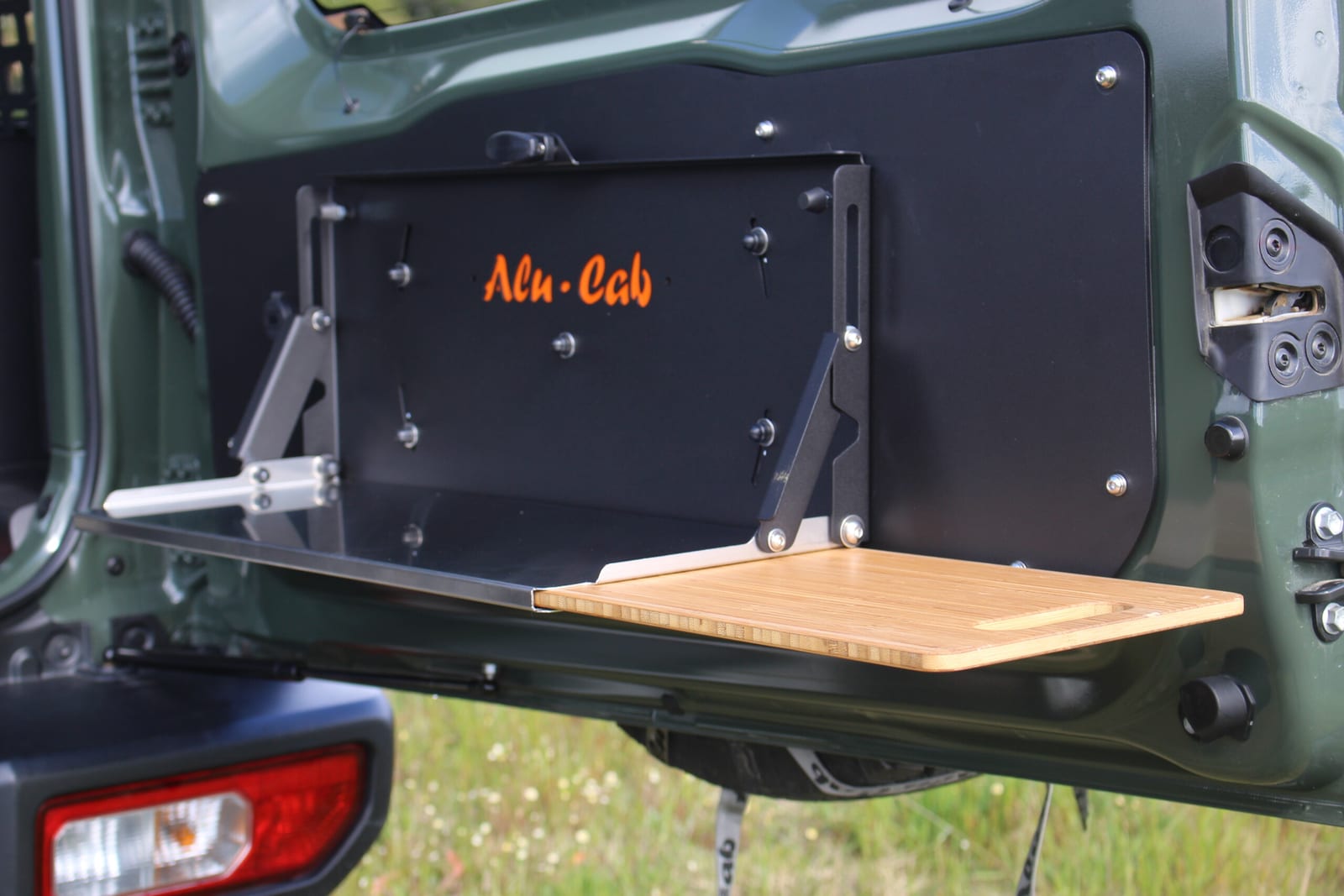 An Alu-Cab drop-down table with a bamboo board folded out on the back door of a green current Suzuki Jimny, grass behind