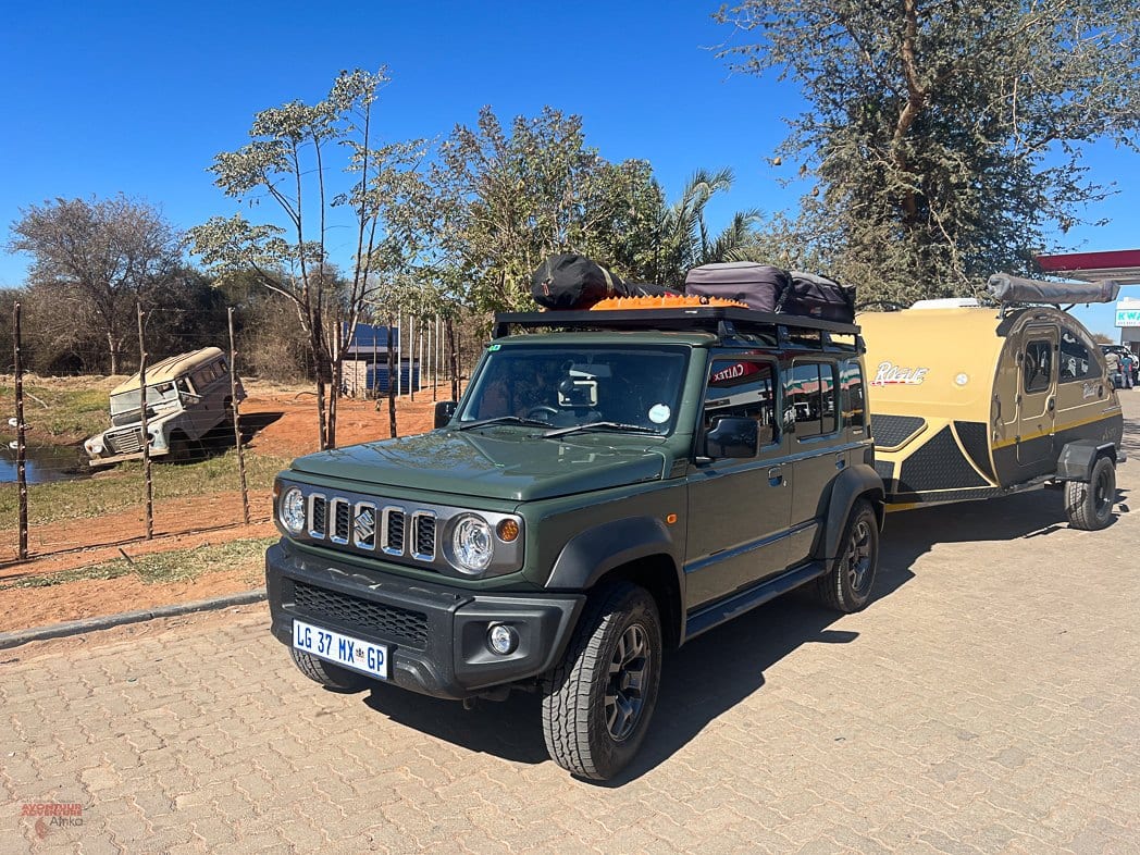 A green Suzuki Jimny 5-door with a loaded roof rack and a teardrop trailer parked at a fuel station in the bushveld