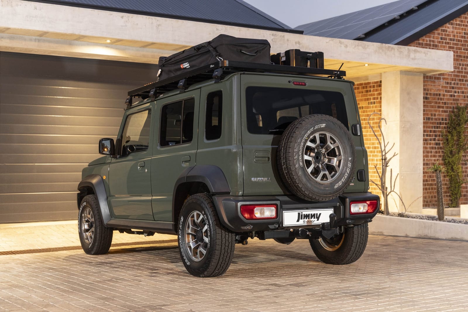 A green Suzuki Jimny 5-door from the rear, parked on a paved driveway, with a roof rack and a standard rear bumper
