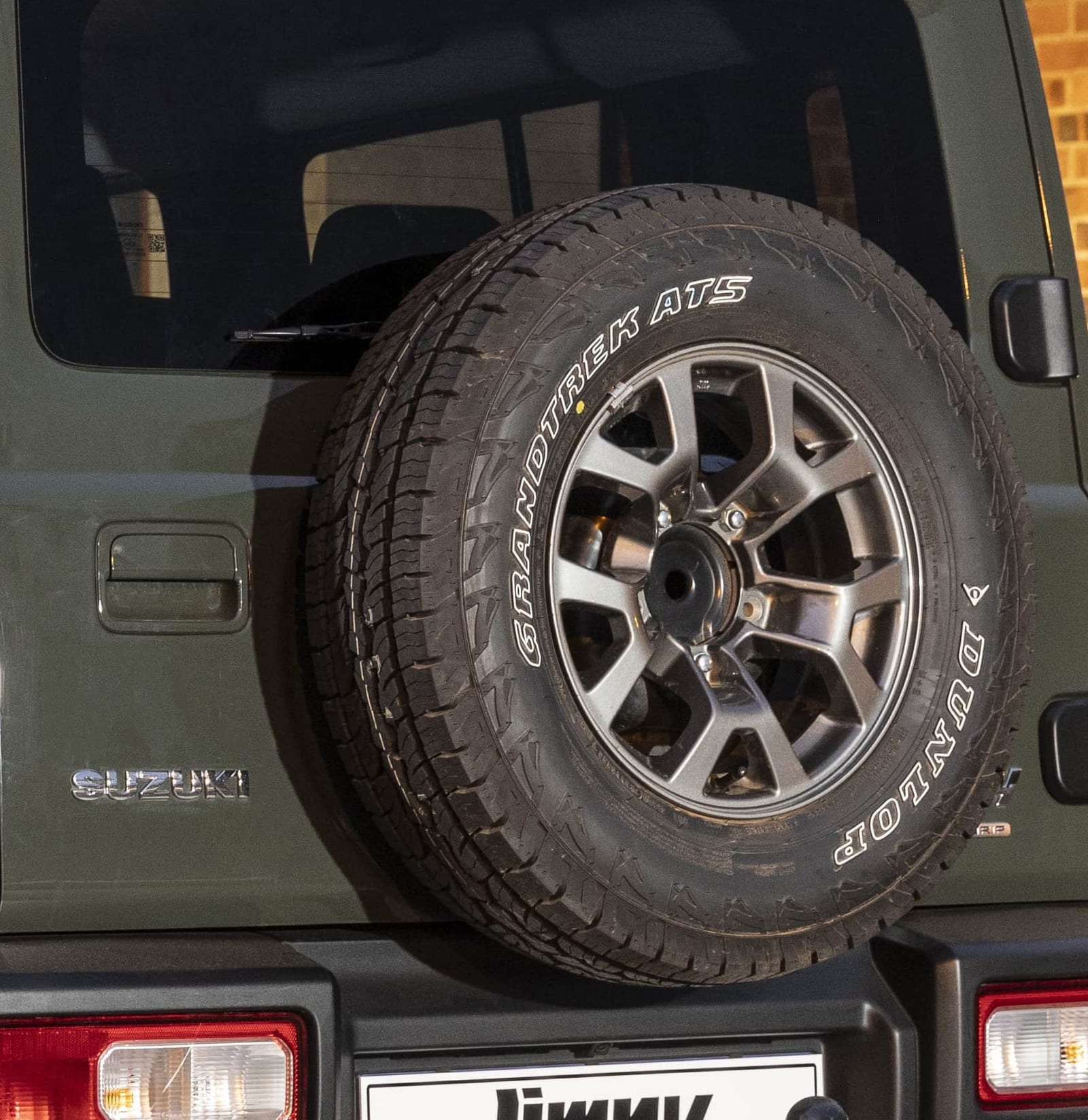 Close-up of the full-size spare wheel on the back door of a green Suzuki Jimny 5-door, on a grey factory alloy with a Dunlop Grandtrek tyre