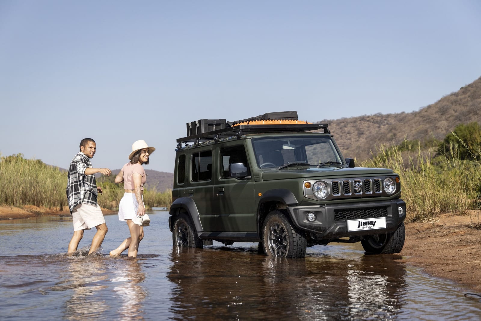 A green Suzuki Jimny 5-door with a roof rack standing in a shallow river on its factory wheels, with two people wading beside it