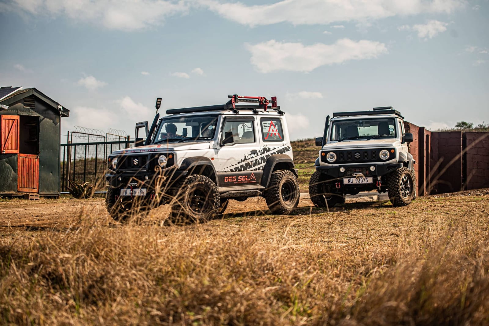 Two white Suzuki Jimny 3-doors, both raised, on black steel wheels and chunky all-terrain tyres, parked on a dry grass farm track