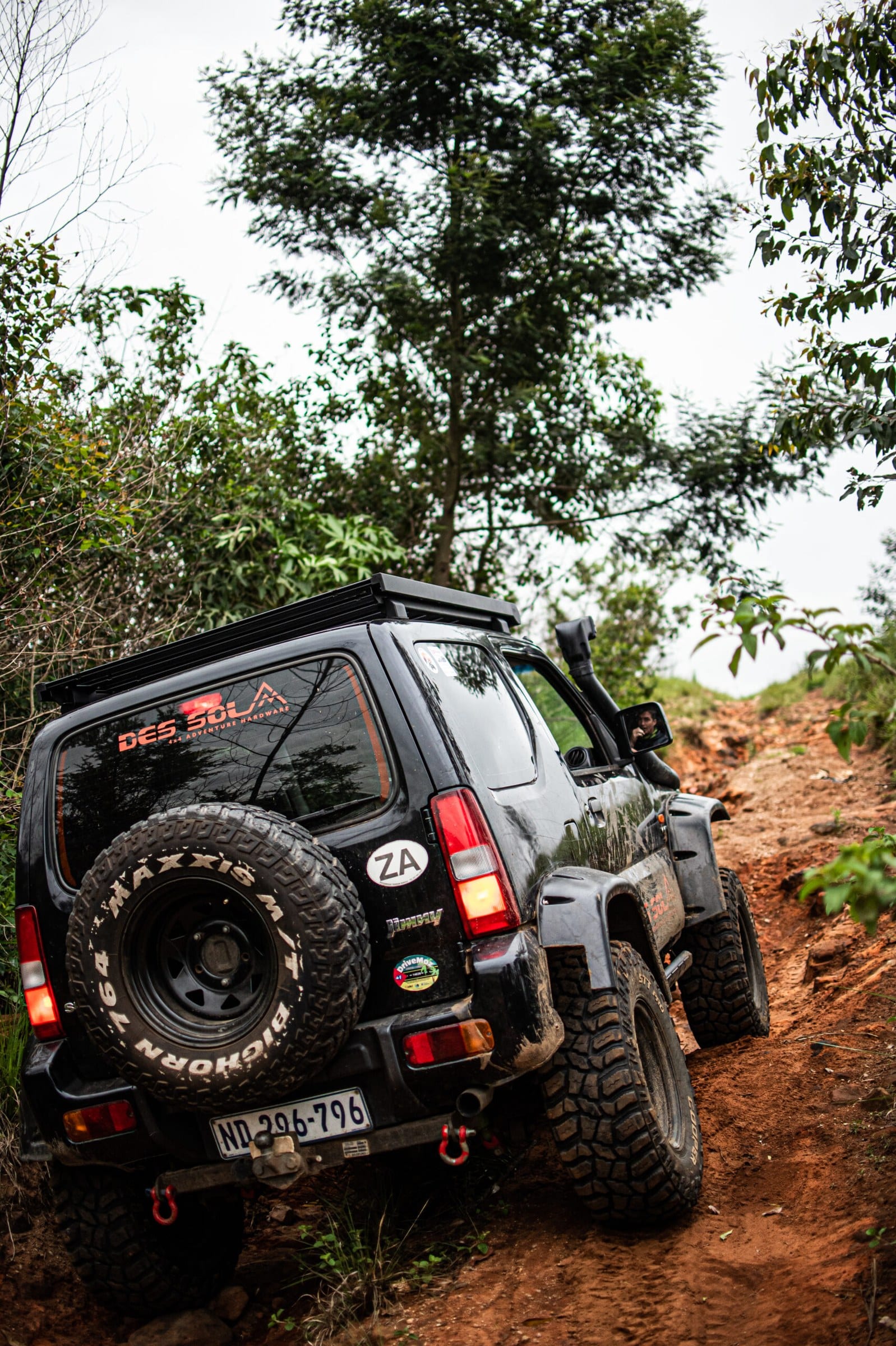 A black older-shape Suzuki Jimny seen from behind, climbing a steep red dirt track through bush, with a chunky spare tyre on the back door