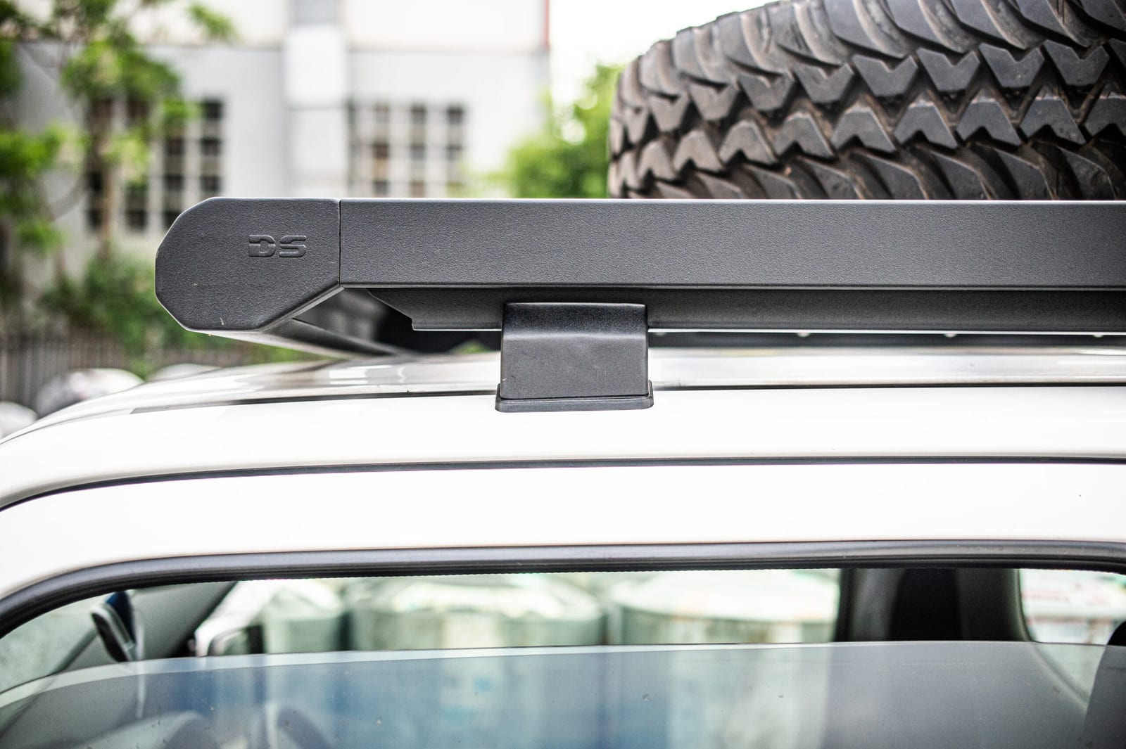 A chunky spare tyre lying flat on a black roof rack above the roof line of a silver Suzuki Jimny