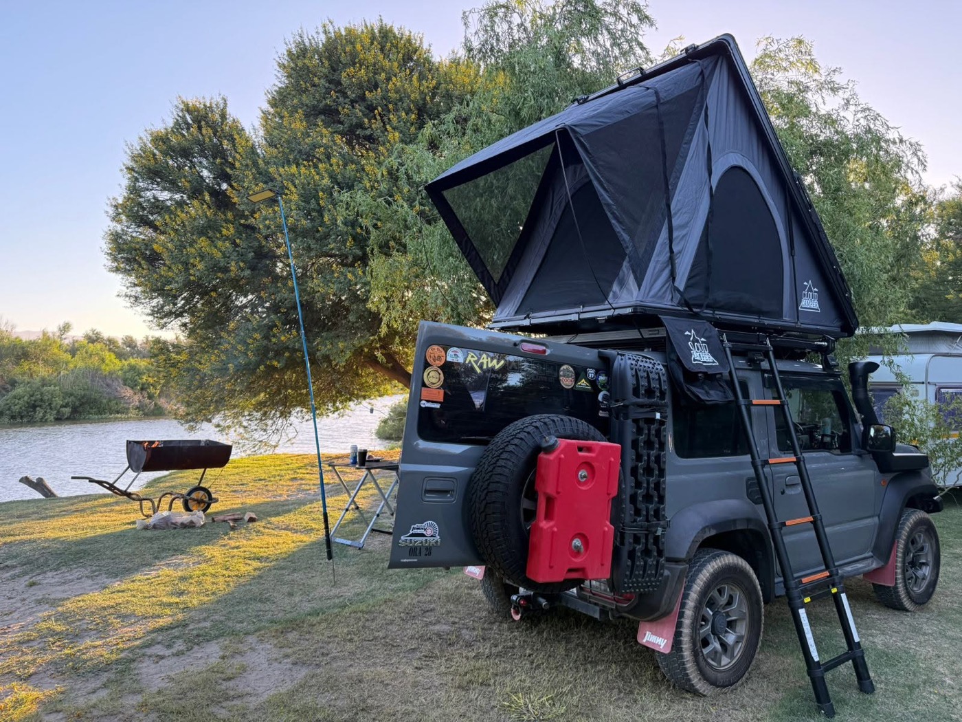 A grey Suzuki Jimny 3-door camped at a riverside with a Cloud Camper wedge tent deployed