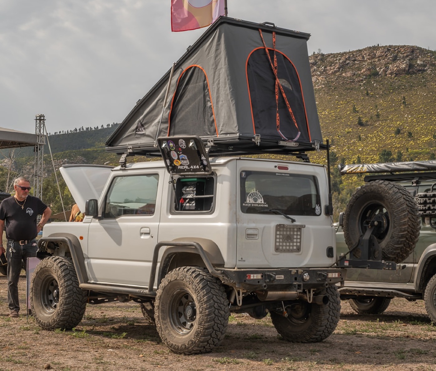 A white 3-door Suzuki Jimny with an Alu-Cab LT-50 wedge tent deployed at a 4x4 gathering