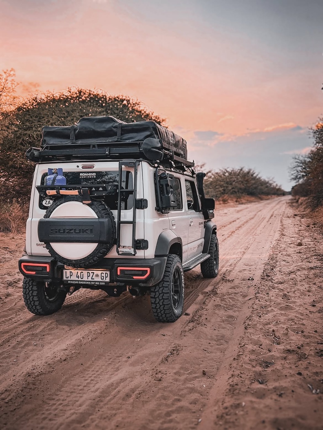 A kitted 5-door Suzuki Jimny driving a Kalahari sand road at sunset with a packed rooftop tent